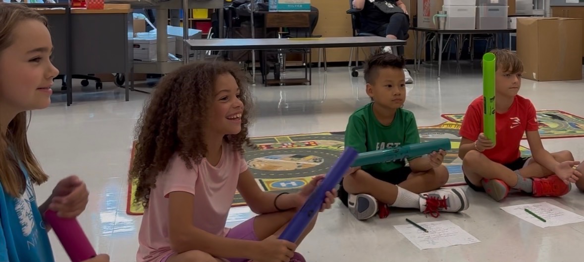 A small group of students gathered in a circle, each with a Boomwhacker, smiling with BoomBot